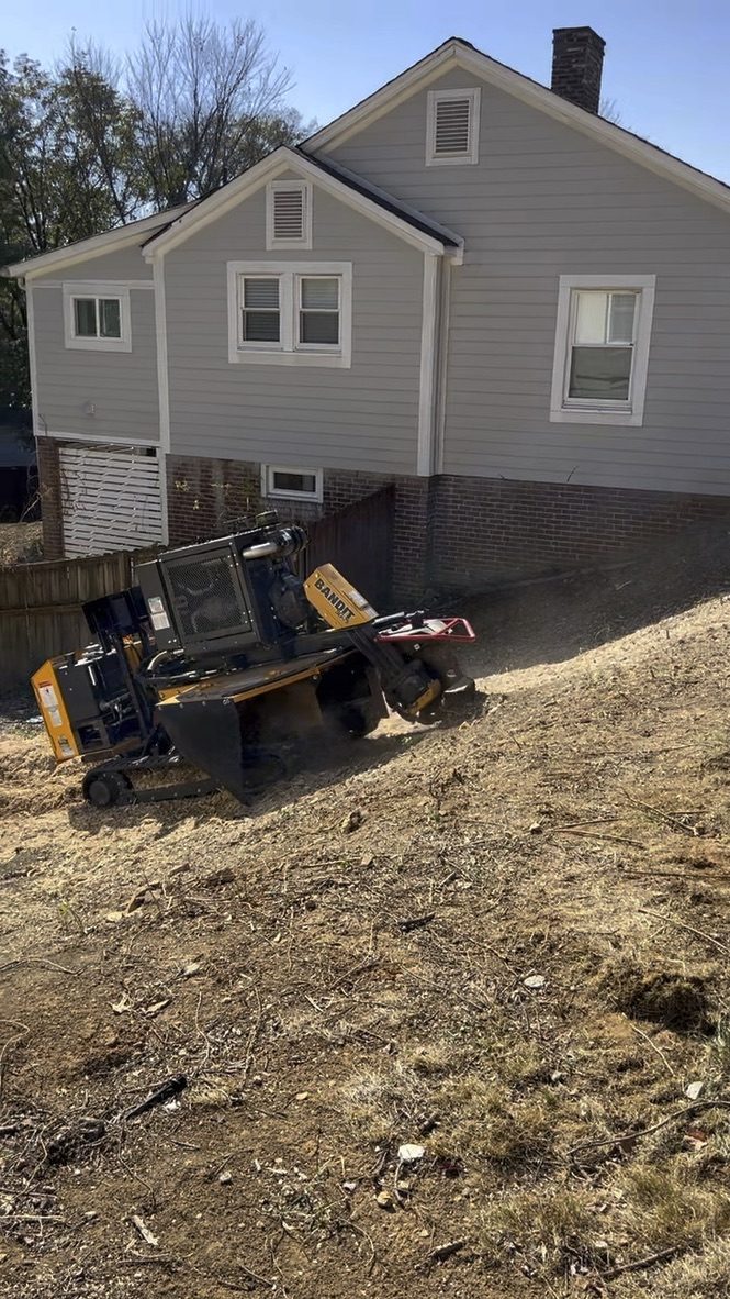 Tracked stump grinder working on a steep dirt slope behind a gray house
